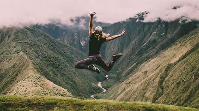 Photo of a woman jumping in the air at the top of a mountain. Other hills and a river are visible below and in the distance.