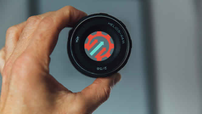 Photo of a man holding the lens of a camera. The geared for growth logo of a gear with upward arrows is in the centre of the lens.