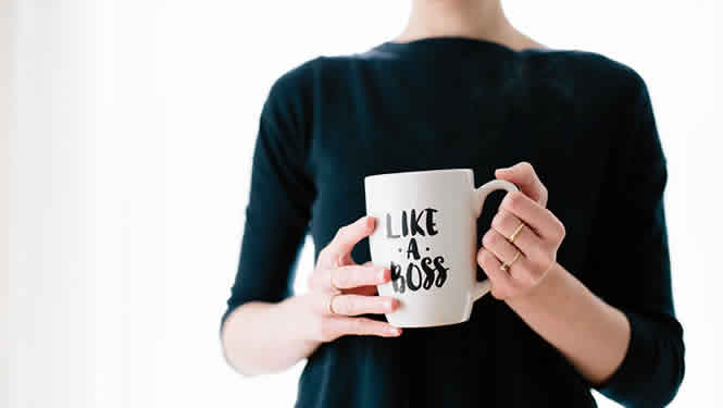 Woman wearing a black top holding a coffee mug with the words 'Like A Boss' on it. She is wearing four gold rings on her fingers.