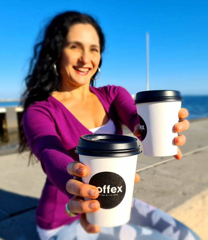 Photo of Josephine Palermo holding out two takeaway coffee cups. She is sitting on a pier with the ocean behind and a clear blue sky.